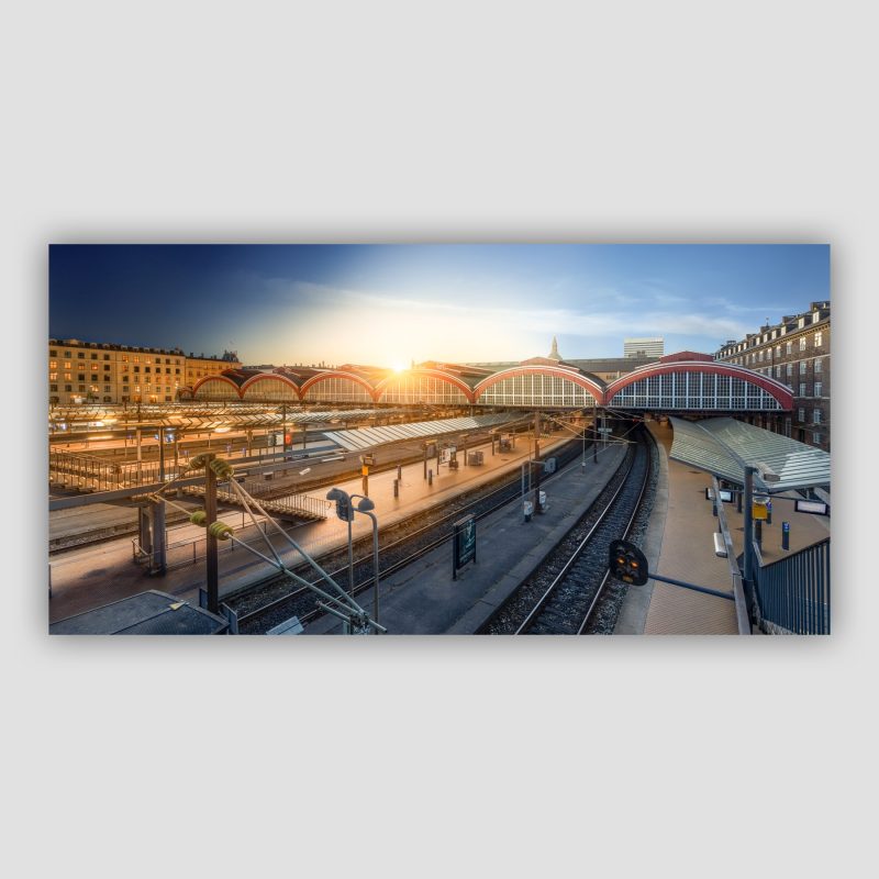 Kunstværket “En dag på Københavns Hovedbanegård” er original fotokunst, der leveres signeret og nummereret i Limited Edition. Københavns Hovedbanegård - pulsen i København. Det er her jyderne kommer ind, her hvor dagen starter og det er her rejsen starter. Her er endnu et "Time blend"-billede, hvor flere billeder taget over et døgn er samlet i ét. En form for timelapse i et enkelt billede. Der er ingen mennesker på billedet – det er et bevidst valg. Det er ikke livet på stederne, vi viser i vores billeder, men snarere de nøgne steder i sig selv.