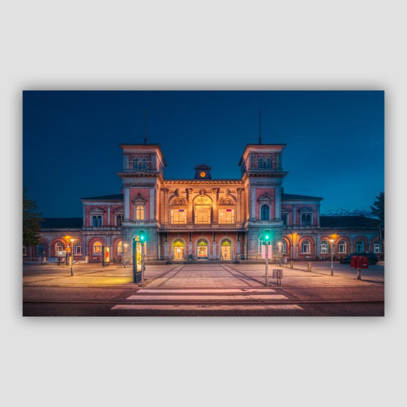 "Aalborg Banegård” er et originalt kunstværk, der leveres signeret og nummereret i Limited Edition. Aalborg Banegård er en af de smukkeste banegårde i Danmark. Den har nogle fælles træk med Danmarks ældste eksisterende banegård, som ligger i Roskilde. Men Aalborgs er bygget mere end 50 år senere og tegnet af en anden arkitekt, nemlig Thomas Arboe. Billedet er taget med fodgængerovergangen i forgrunden på en fin aften.