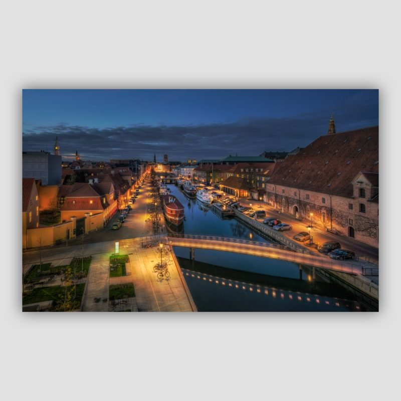 “Stille aften ved Frederiksholms Kanal” er et originalt kunstværk, der leveres signeret og nummereret i Limited Edition. Det her billede af Frederiksholm Kanal har været på ambolten mange gange, men nu blev det endelig færdigt! Når det snerper til, så må man jo træffe nogle bindende beslutninger 😅 Vi har flere billedserier af samme motiv og har derfor kunnet vælge hvilken historie billedet skulle fortælle. I lang tid var idéen at inkludere de mange lysstriber fra bilerne der susede rundt, men nu i forbindelse med, at vi lægger sidste hånd på billederne til vores bog ombestemte vi os. Det er sådanne steder, det godt at have en partner in crime, fordi vi ofte træffer de kunstneriske beslutninger i fælleskab. Resultatet er et billede, som har meget mere ro og giver plads til, at man kan gå på opdagelse i de mange detaljer uden hektiske bilstriber. Og så passer det meget bedre til vores mennesketomme billeder.