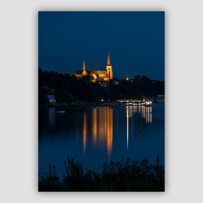 “Double Reflection of Roskilde Cathedral” er et originalt kunstværk, der leveres signeret og nummereret i Limited Edition. Roskilde Domkirke knejser på toppen af bakken i Roskilde og spejler i fjordens vand i den dybe blå time. Der er lys i en pavilion på en bådebro og spejlingen af den har næsten samme facon som spejlingen af kirken.